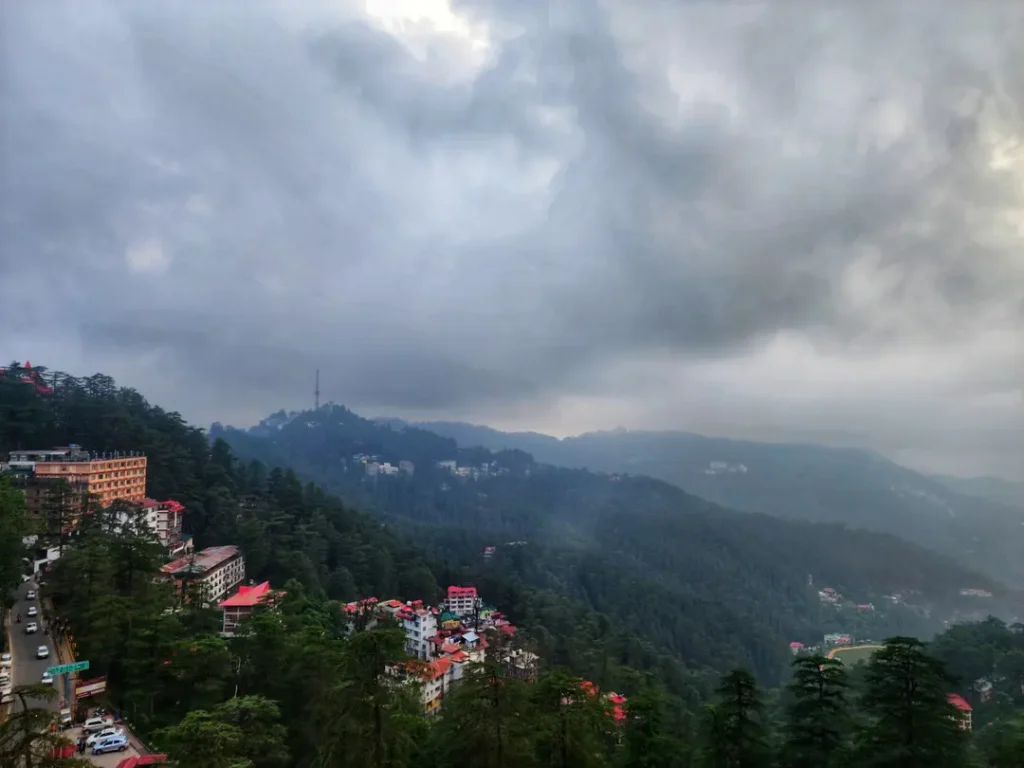 Scenic panoramic view of Shimla's lush green hills, pine forests, colorful buildings, and dramatic monsoon clouds as seen from Hotel Surya Shimla near Mall Road.