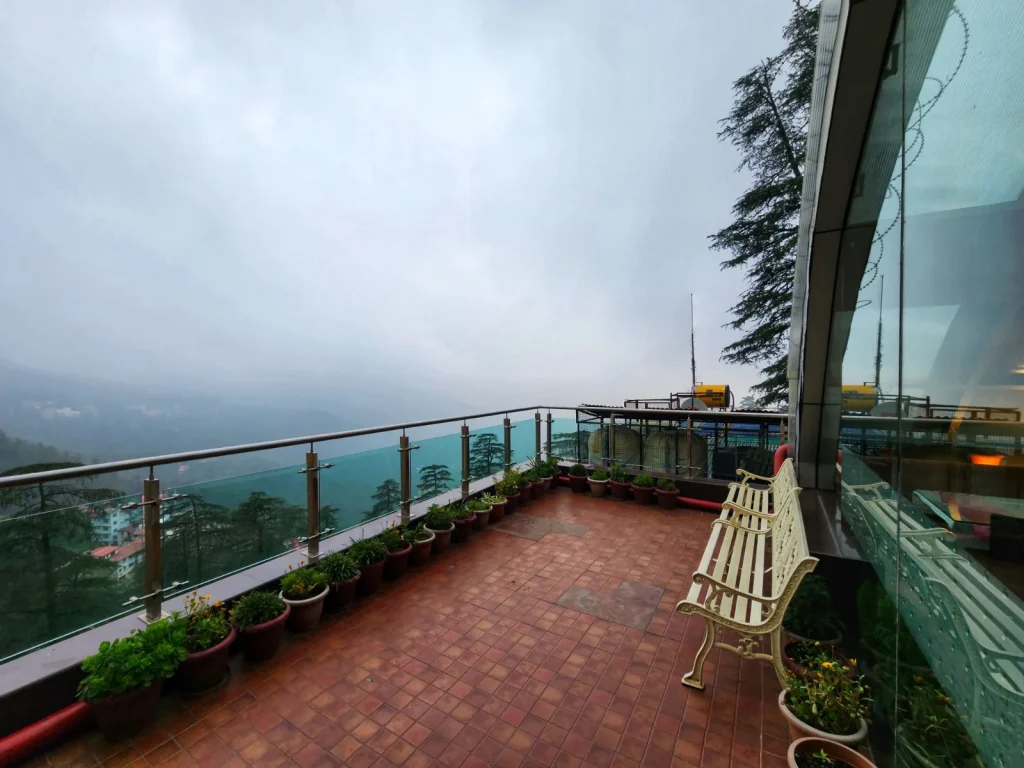 Open terrace at Hotel Surya Shimla featuring glass railings, comfortable seating, potted plants, and breathtaking panoramic views of the Himalayan mountains and lush valleys.